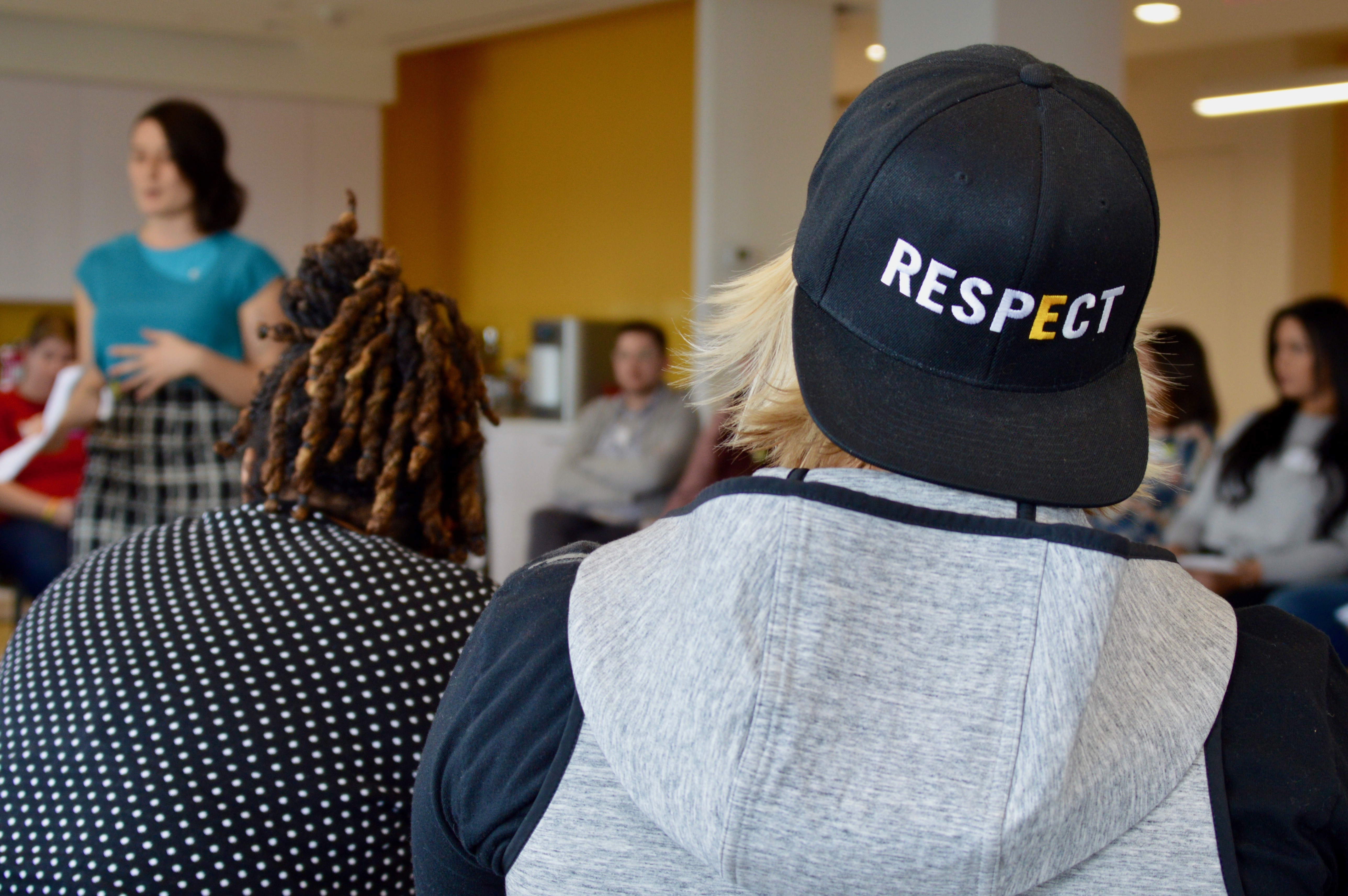A blond adult volunteer with their back to the camera attends the 2019 NYC Training of Trainers. They wear a GLSEN black snapback with the text: RESPECT. They sit with several other volunteers in a U-shape of chairs listening to a standing GLSEN National staff member in a blue shirt. 