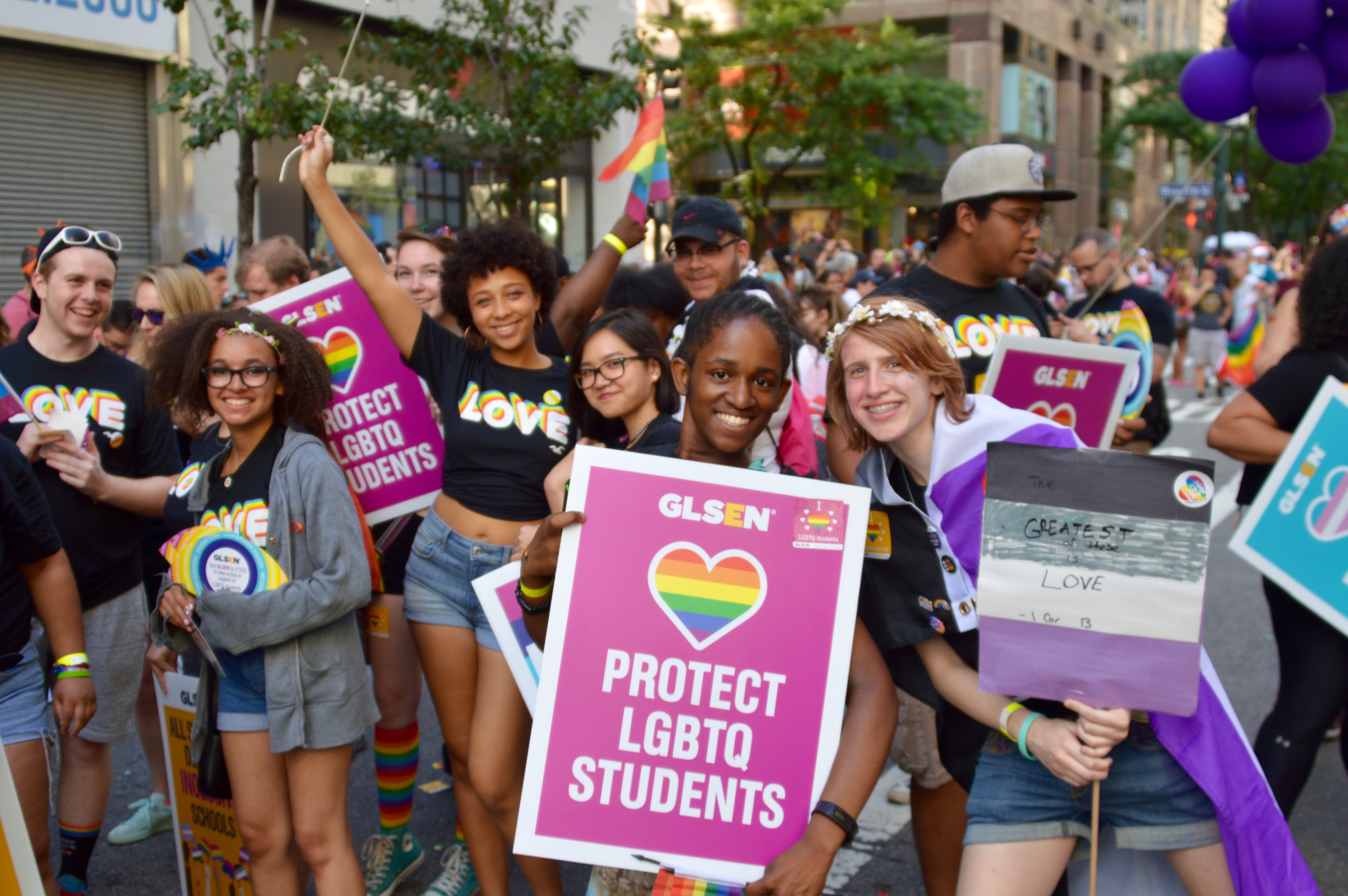A group of student vounteers smile at the camera while marching in the 2018 NYC Pride March. They wear GLSEN Love shirts and hold up GLSEN posters that read: Protect LGBTQ Students. 