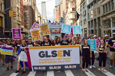 A large group of GLSEN volunteers, all wearing black tshirts that say LOVE, holds up posters and a banner as they march in the 2018 NYC Pride Parade. A double-decker bus behind them also has a GLSEN banner and more volunteers on the roof. The banners reads: GLSEN, Championing LGBT Issues in K-12 Schools Since 1990; GLSEN, Creating LGBTQ Inclusive Schools. The posters read: Resist; Protect LGBTQ Students; All Students Deserve Inclusive Schools; Protect Trans Students.