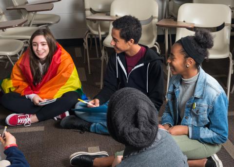 A group of students sit in a circle on the floor of a classroom with the desks pushed aside. They smile and hold notebooks. The first student on the left is wrapped in a rainbow flag. 