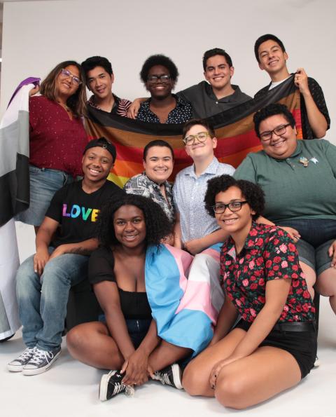 Image is a group of queer students holding the rainbow flag, the trans flag, and the asexual flag.