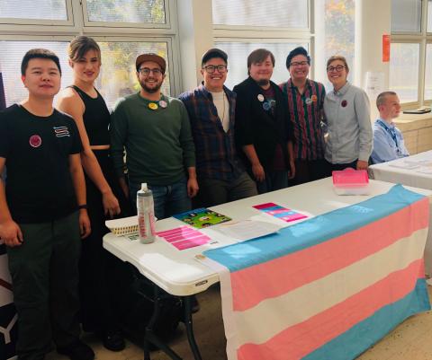 A group of educators stands behind a table that has the trans flag on the front of it.