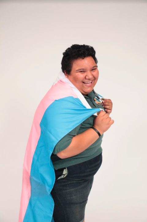 Reggie standing to the side with a trans flag draped over his shoulders. 
