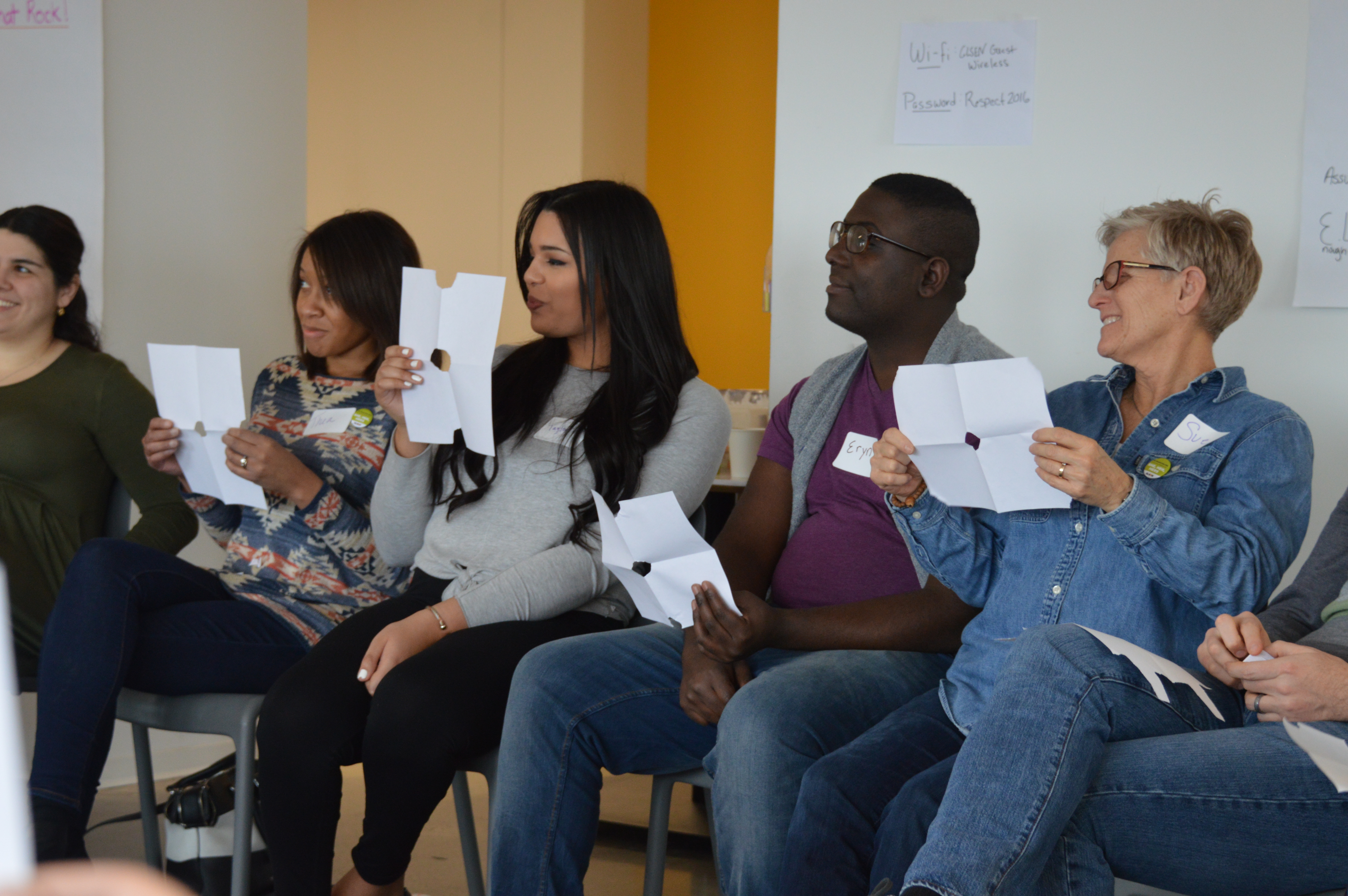 Four sitting adult volunteers participate in an activity at GLSEN's NYC Training of Trainers, or TOT. They smile and hold up pieces of white paper with fold lines and a circle cut in the center. 