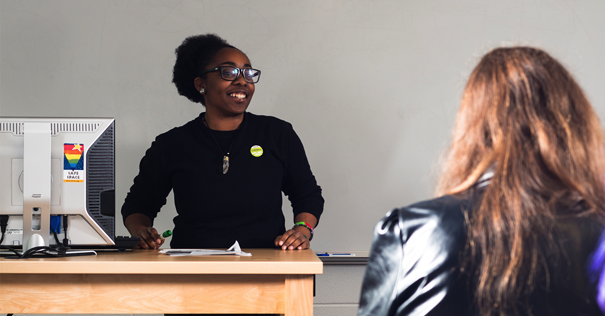 A teacher in front of her classroom. She is wearing a pronoun button and her computer has a Safe Space Sticker on it.