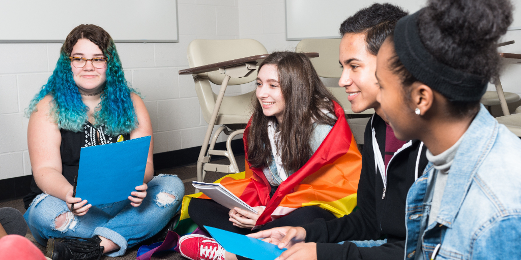 Students sitting in a circle in a classroom. One is wearing a rainbow flag.
