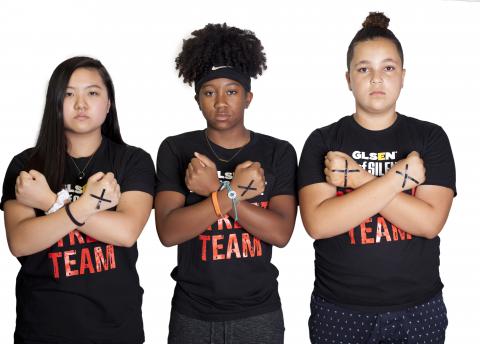 Three National Student Council members, all members of GLSEN's Day of Silence Street Team, stand in a row and hold their arms up to their chest in an X. On the backs of their hands are X's in black tape. 