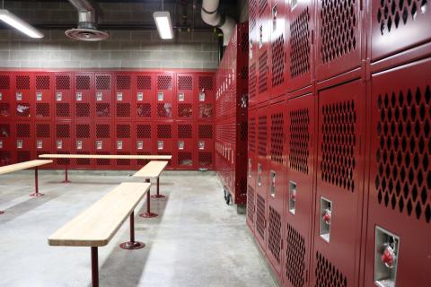 This is an empty school locker room with four wooden benches and two banks of red lockers at a 90 degree angle from each other. 