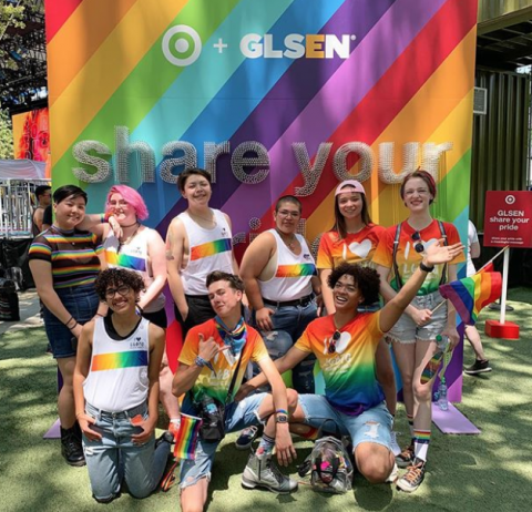 A group of student volunteers, including several National Student Council members, smile and pose in front of a rainbow board at GLSEN's 2019 Share Your Pride event partnered with Target. The students on the left are wearing a white tank-top with a rainbow gradient band and the small text: I Heart LGBTQ. The students on the right are wearing a rainbow gradient t-shirt with the larger text: I Heart LGBTQ.