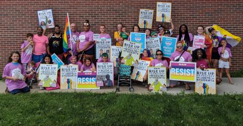A large group of volunteers and students from GLSEN Omaha smile and hold up rainbow flags and illustrated Rise Up posters. The posters read: rise up to protect immigrant students; rise up to protect Muslim students; rise upt o protect LGBTQ students; RESIST; rise up to protect trans and GNC students; rise up to protect black students; protect trans students; let youth lead.