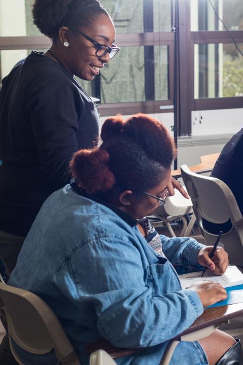 A smiling young educator in a classroom stands over a high school student's desk and points at their notebook.