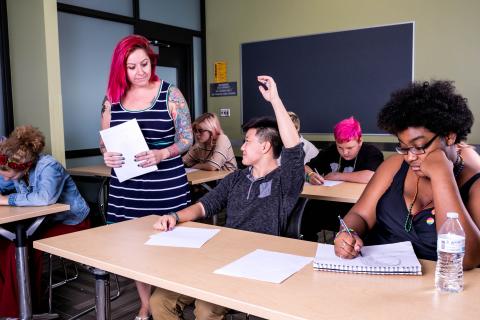 Four classroom tables of students write in their notebooks. A pink-haired educator holding papers approaches a student with their hand raised.