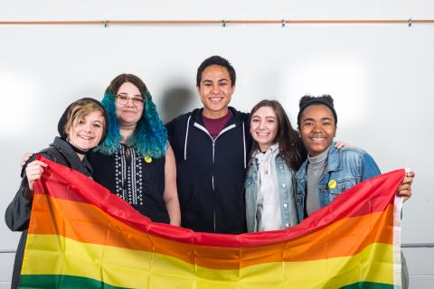A group of five high school students hold up a rainbow flag and smile at the camera. Two students are wearing GLSEN pronoun pins. 