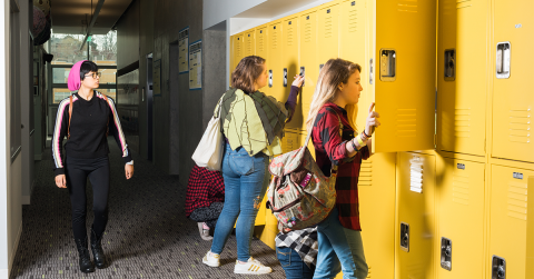 A student wearing a pink hat walks past a bank of bright yellow lockers. At the lockers are four other students, two kneeling, who are opening their lockers or rummaging inside. 