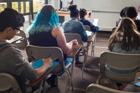 Seven high school students in a classroom sit working at their desks with their backs to the camera. 