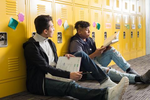 Two high school students sit with their legs stretched out against a bank of yellow lockers with post-it notes with names stuck to them. The students are studying their textbooks. One looks over at something on the other's book. 