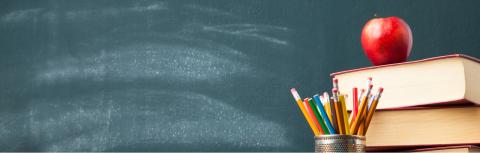 A stack of books, with a cup of pencils and a red apple in front of a blackboard.