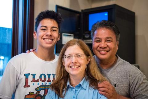 Three humans stand together smiling at the camera. The person on the right has his hands on each persons shoulders. 