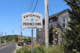 Road sign that reads "Entering Provincetown". There is a brown house on the left behind the sign and a road on the right. The sky is blue with no clouds.
