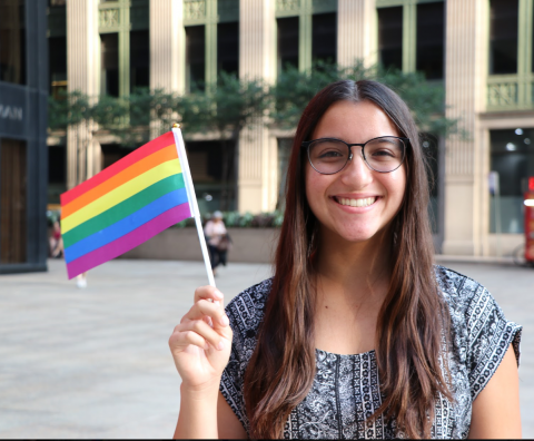 A female-presenting Latinx student in glasses smiling and holding up a rainbow flag.