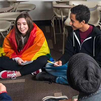 A group of students at a GSA meeting sitting on the floor. One student is wearing a rainbow flag.