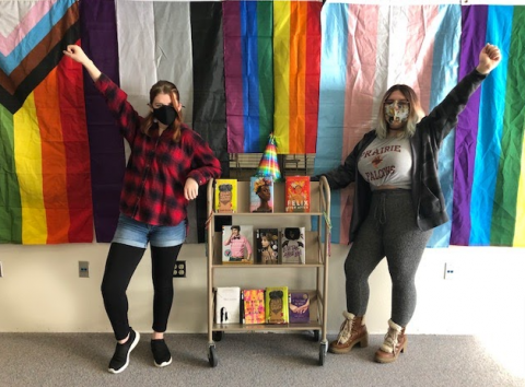 Two individuals with masks with their fists up showcasing LGBTQ+ books. There are various LGBTQ+ representing flags behind them